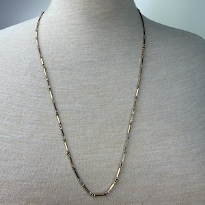 May include: A gold-colored necklace with a delicate design. The necklace features small, rectangular links connected by thin, circular rings. The necklace is displayed on a mannequin, showcasing its length and style.