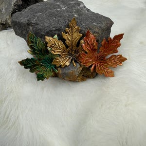 May include: Three decorative maple leaf ornaments in metallic finishes of green, gold, and copper. Each leaf features detailed veining and a textured surface, arranged in a cluster against a dark, jagged rock formation.