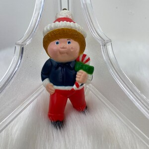 May include: A vintage Christmas ornament featuring a Cabbage Patch Kid doll wearing a blue jacket, red pants, and a white and red Santa hat. The doll is holding a candy cane.