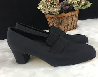 May include: Black heeled loafers with a buckle detail. The shoes are made of a soft fabric and have a low block heel.
