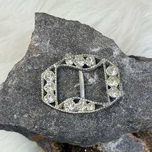 May include: A silver-toned metal buckle with a decorative design featuring a row of clear rhinestones around the perimeter. The buckle is designed to be used for a belt or strap.