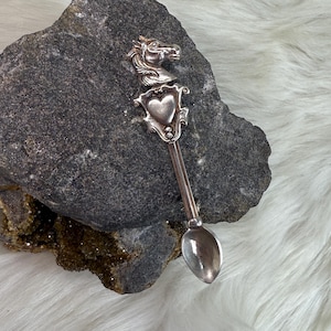 May include: Silver spoon with a horse head and heart design. The spoon is on a gray rock.