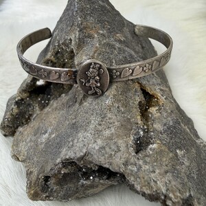 May include: Silver-toned cuff bracelet featuring a central circular charm with a Minnie Mouse design. The bracelet has a heart and floral pattern along the band. The bracelet is displayed on a gray rock.