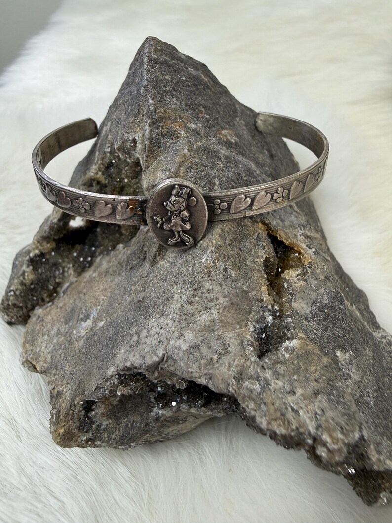 May include: Silver-toned cuff bracelet featuring a central circular charm with a Minnie Mouse design. The bracelet has a heart and floral pattern along the band. The bracelet is displayed on a gray rock.