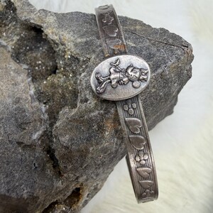 May include: A silver-toned bracelet featuring an oval centerpiece with a cartoon character design. The band is decorated with heart and flower patterns. The bracelet is displayed on a gray rock surface.