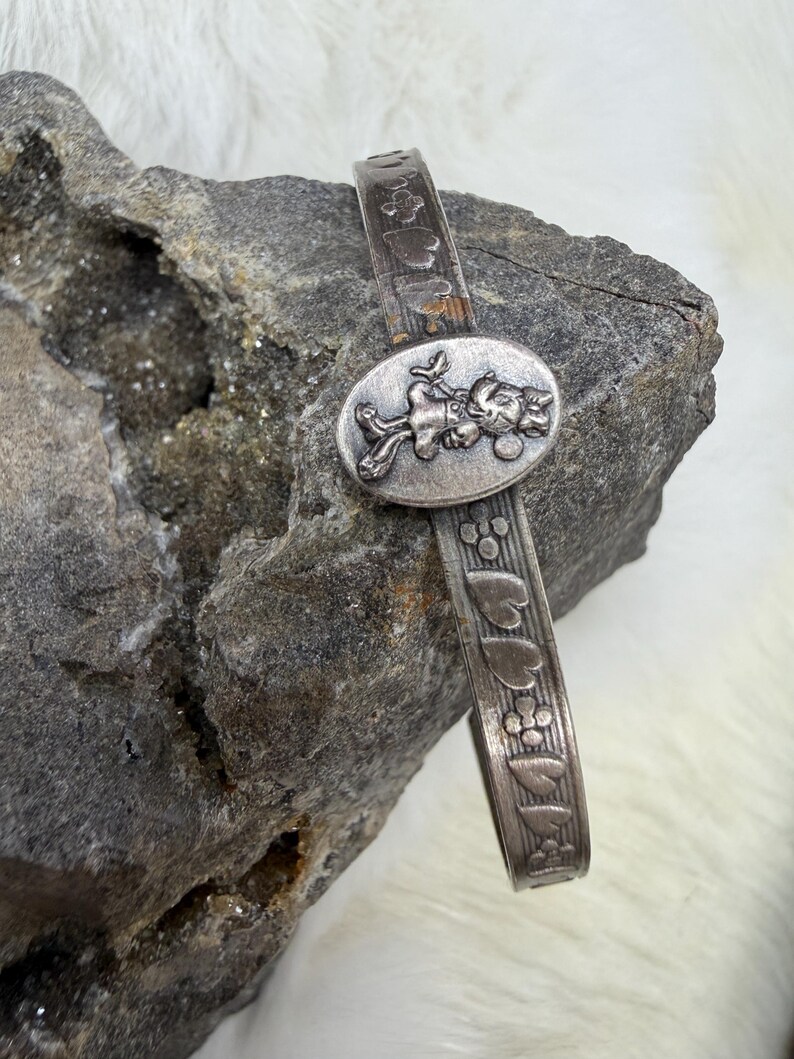 May include: A silver-toned bracelet featuring an oval centerpiece with a cartoon character design. The band is decorated with heart and flower patterns. The bracelet is displayed on a gray rock surface.