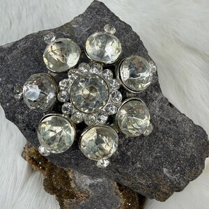 May include: A silver-toned brooch with a starburst design. The brooch features a large, central clear crystal surrounded by smaller crystals and six large, faceted, clear crystal orbs. The brooch is set on a dark gray stone.