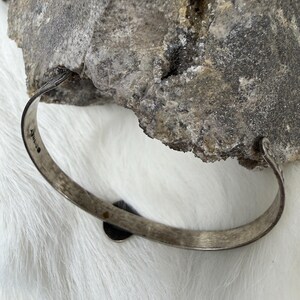 May include: A silver-toned bangle bracelet with a small, dark circular accent. The bracelet has a simple, slightly textured design. The background features a rough, gray stone and white fur.