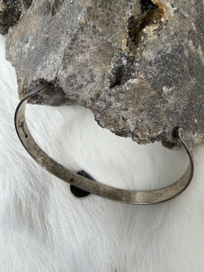 May include: A silver-toned bangle bracelet with a small, dark circular accent. The bracelet has a simple, slightly textured design. The background features a rough, gray stone and white fur.