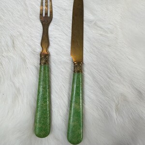 May include: A vintage fork and knife set. The fork and knife have brass blades and tines, with green handles. The handles have a textured appearance. The set is displayed on a white, fluffy surface.