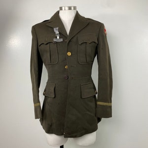 May include: A vintage military jacket in olive green with gold buttons and a red and white patch on the right sleeve. The jacket has a double breasted closure and two large pockets on the front.
