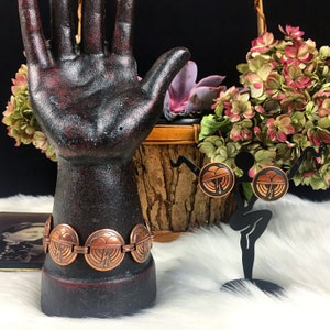 May include: A copper bracelet with a geometric design of overlapping hearts. The bracelet is on a black hand-shaped display stand. Two copper earrings with the same heart design are on a black stand with a white fluffy surface.
