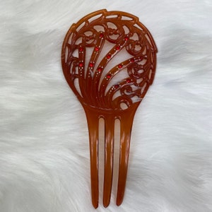 May include: A brown plastic hair comb with a decorative, ornate design. The comb has three tines and features a circular, openwork design with red rhinestones.