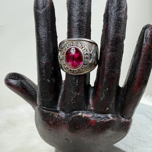 May include: A silver-toned ring with a large, round, ruby-red stone. The ring has detailed engravings around the band, including text and symbols. The ring is displayed on a dark, textured hand-shaped stand.