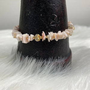 May include: A delicate bracelet made of small, irregular pieces of white and pink seashells, interspersed with tiny gold beads. The bracelet is displayed on a dark brown surface.
