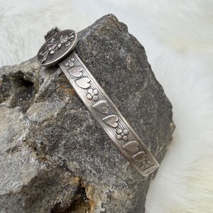 May include: A silver-toned metal bracelet with a floral and heart design. The bracelet features a circular clasp with a detailed insect design. The bracelet is resting on a gray rock.