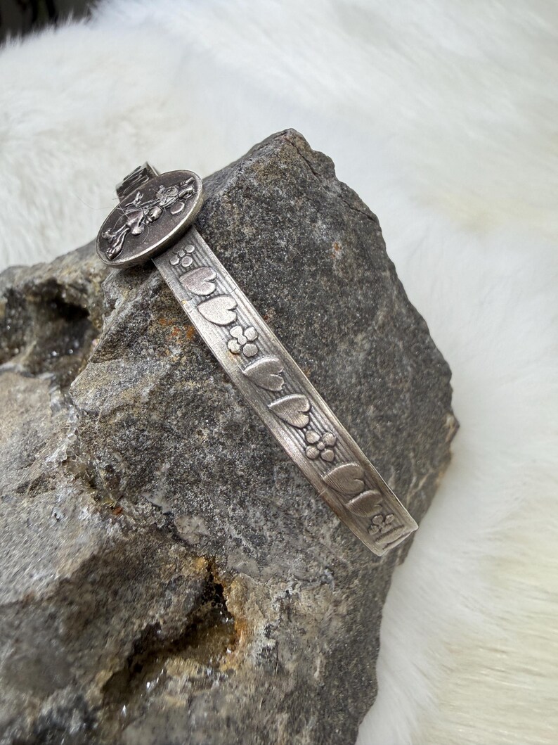 May include: A silver-toned metal bracelet with a floral and heart design. The bracelet features a circular clasp with a detailed insect design. The bracelet is resting on a gray rock.
