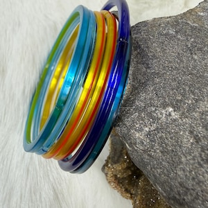 May include: A stack of colorful bangles in shades of blue, green, yellow, and red. The bracelets are made of a translucent material and are stacked together. The bangles are resting on a white, fluffy surface next to a gray rock.