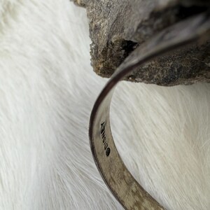 May include: A silver-toned bracelet with the Disney logo stamped on the inside. The bracelet is curved and sits against a white, fluffy surface and a gray rock. The bracelet has a textured surface.