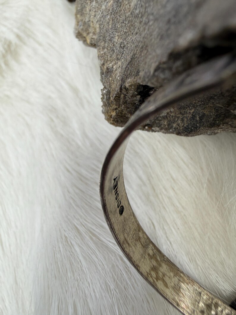 May include: A silver-toned bracelet with the Disney logo stamped on the inside. The bracelet is curved and sits against a white, fluffy surface and a gray rock. The bracelet has a textured surface.