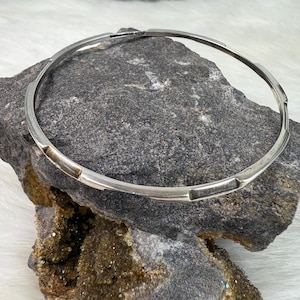 May include: A silver bangle bracelet with a geometric design. The bracelet is made of a thick, solid band of silver and has a simple, modern look.