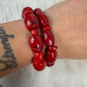 May include: Two strands of bright red beaded bracelets. The beads are various shapes and sizes, including round and oblong. The bracelets are worn on a wrist, with a tattoo visible on the arm.