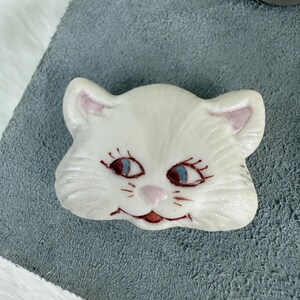 May include: A white cat-shaped brooch with blue eyes, pink ears, and a pink nose. The cat has a smiling expression and is set against a gray background. The brooch is likely made of ceramic or a similar material.