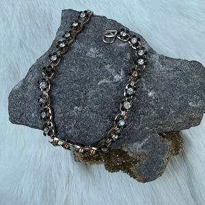 May include: A vintage necklace with a gold-tone chain and black flower accents. Each flower features a clear, round rhinestone center. The necklace is displayed on a gray stone surface.