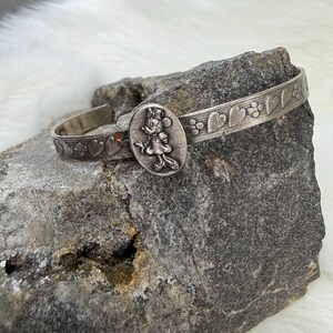 May include: Silver-toned cuff bracelet featuring a Minnie Mouse cameo. The bracelet has a heart and floral pattern along the band. The oval cameo shows Minnie Mouse in a dress and bow. A charming piece of jewelry.