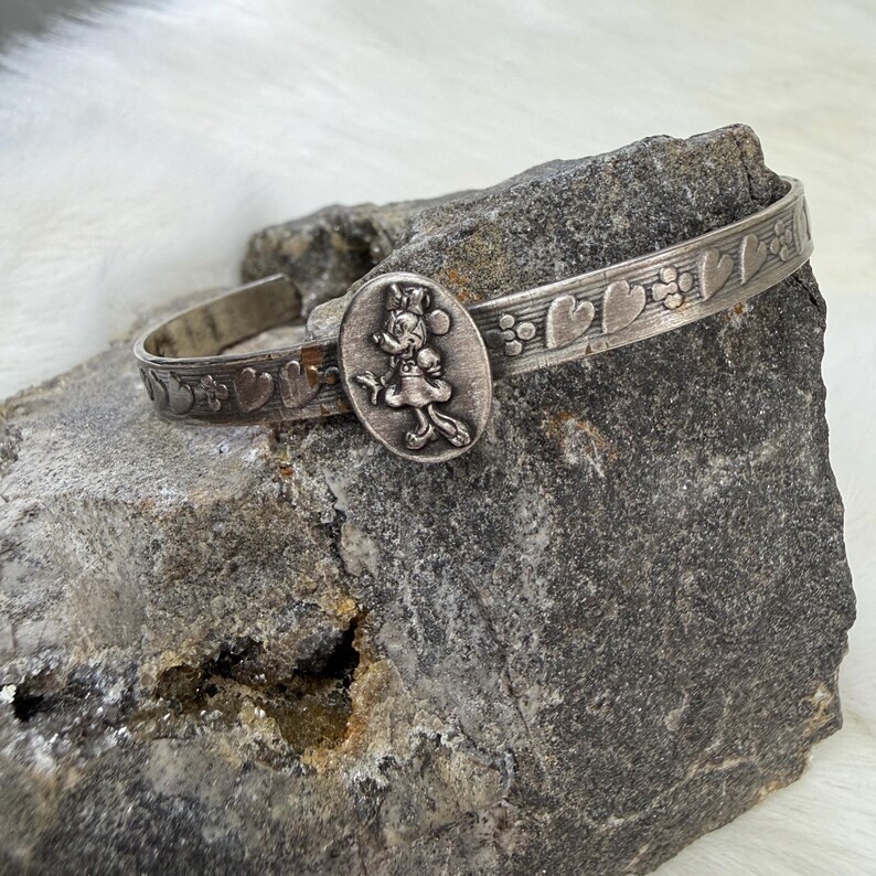 May include: Silver-toned cuff bracelet featuring a Minnie Mouse cameo. The bracelet has a heart and floral pattern along the band. The oval cameo shows Minnie Mouse in a dress and bow. A charming piece of jewelry.