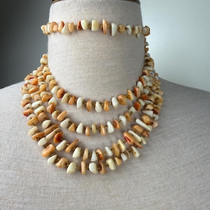 May include: A multi-strand necklace and matching bracelet set. The jewelry features irregularly shaped, polished beads in shades of cream, orange, and tan. The beads are strung on a gold-colored wire, creating a layered effect. The necklace is displayed on a mannequin.