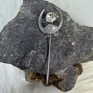 May include: A silver-toned hat pin with a crescent-shaped top, a large clear gemstone, and a diamond-shaped accent. The pin has a long, slender needle. The pin is likely vintage or antique.