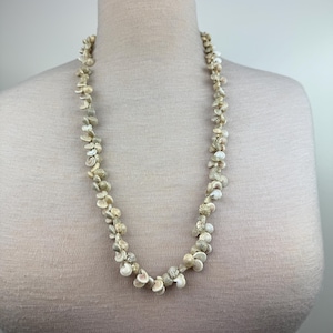 May include: A necklace made of small, natural seashells in shades of cream, beige, and off-white. The shells are strung together to create a long, flowing strand. The necklace is displayed on a mannequin.