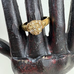 May include: A gold-toned ring with a cluster of small, clear crystals forming a rounded design. The ring is set on a dark, textured surface, showcasing its intricate details and sparkling appearance. The ring's band is a simple, polished gold.