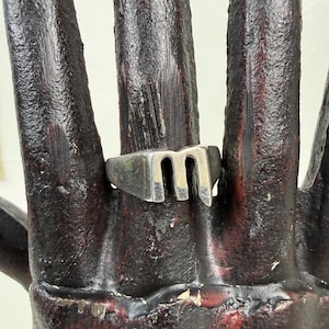 May include: A silver-toned ring with a capital letter "M" design. The ring has a rectangular band and a raised, stylized "M" shape. The ring is worn on a dark, textured surface.