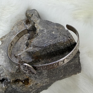 May include: A silver-toned cuff bracelet with a decorative design of hearts and flowers. The bracelet is resting on a gray rock surface. The bracelet has a small, round clasp with a floral design.