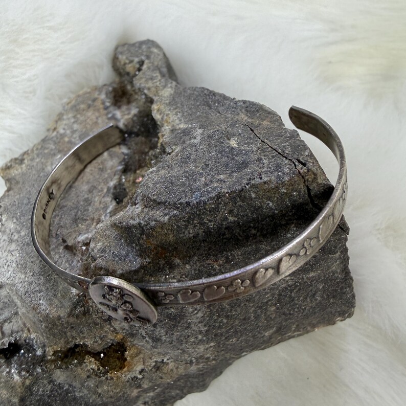 May include: A silver-toned cuff bracelet with a decorative design of hearts and flowers. The bracelet is resting on a gray rock surface. The bracelet has a small, round clasp with a floral design.