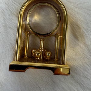 May include: A gold-colored, arched-top decorative clock with a clear, circular center. The clock features a pendulum with three small, gold spheres. The base is rectangular and also gold.