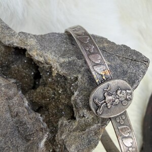 May include: A silver-toned bracelet with an oval centerpiece featuring a cartoon character. The band is decorated with embossed hearts and flowers. The bracelet is resting on a gray rock.