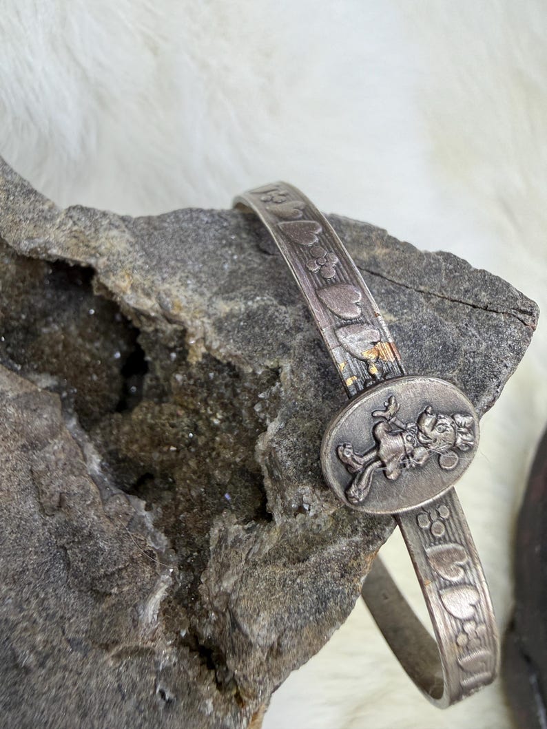 May include: A silver-toned bracelet with an oval centerpiece featuring a cartoon character. The band is decorated with embossed hearts and flowers. The bracelet is resting on a gray rock.