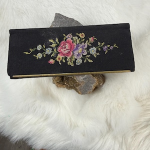 May include: A black clutch with a floral cross-stitch design. The embroidered flowers feature pink, purple, and yellow hues. The clutch has a gold-colored frame and clasp, and is resting on a rock.