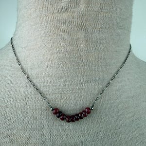 May include: A delicate necklace featuring a series of small, faceted, deep red gemstones. The gemstones are strung on a fine, dark metal chain. The necklace is displayed against a neutral background, highlighting the jewelry's color and design.