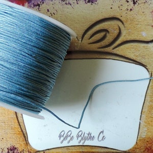 May include: A spool of light blue waxed cord, wound around a white plastic bobbin. The cord is unwinding and trailing off to the right.
