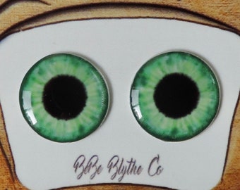 Puede incluir: Placa de madera con dos cabujones de ojos verdes con pupilas negras, sobre un fondo blanco. El fondo es marrón claro con un diseño de lazo y el texto "Bbze Blythe Co". La placa tiene salpicaduras de pintura coloridas.