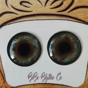 May include: Two green and brown doll eyes with black pupils, displayed on a white card with the text "Blythe Blythe Co.", against a brown background with colorful paint splatters.