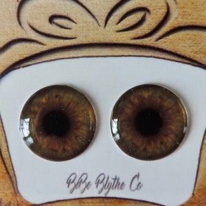 May include: Two brown and gold doll eyes with black pupils, displayed on a white card with the text "Bye Bye Blythe Co.". The card is on a brown background with colourful paint splatters.