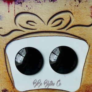 Puede incluir: Dos botones redondos negros con una superficie reflectante se muestran en una caja de regalo blanca con un lazo marrón en la parte superior. La caja está sobre un fondo marrón con salpicaduras de pintura de colores. El texto "B&B Blythe Co" está impreso en la parte inferior de la caja.