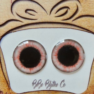 May include: Two pink and white doll eyes with black pupils, displayed on a white background with a brown ribbon and the text "B&B Blythe Co."