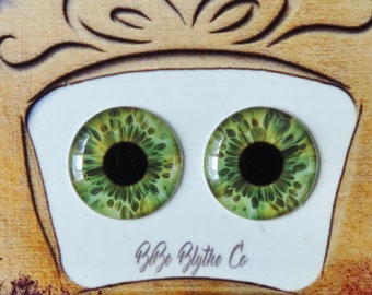 Puede incluir: Un par de ojos de muñeca Blythe verdes con pupilas negras, presentados en una tarjeta blanca con el texto "B&B Blythe Co.". La tarjeta está colocada sobre un fondo marrón con salpicaduras de pintura roja, naranja, amarilla, verde, azul y morada.
