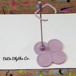 May include: A lavender-colored doll stand with a four-leaf clover base and a metal rod. The stand has a curved top for holding a doll. The text "BeBe Blythe Co." is printed on a white card behind the stand.